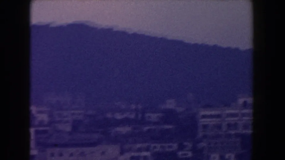 1960 Italy 8mm frame showing low-rise urban buildings beneath a dark hillside, with pronounced blue-purple color shift.