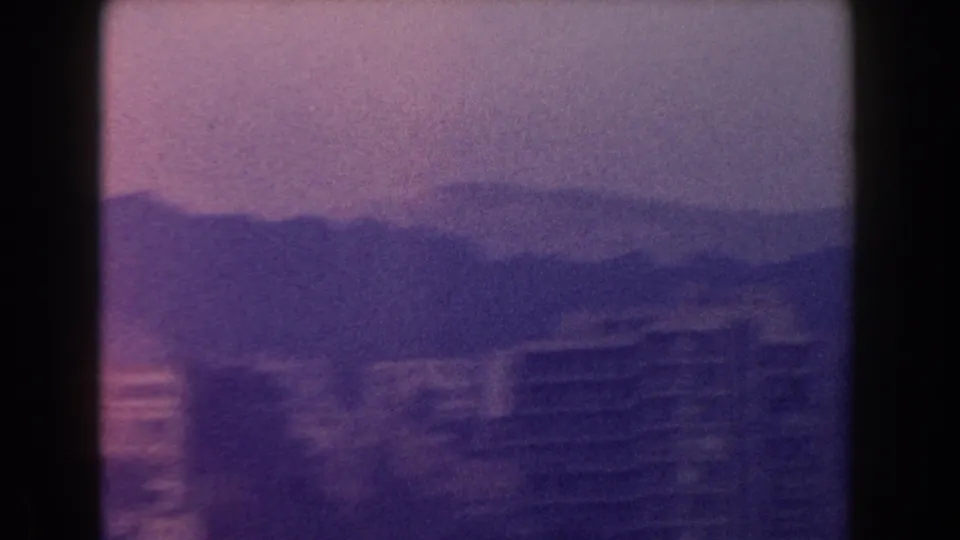 1960 Italy 8mm view of distant buildings, including a larger block, with hills fading into haze behind them.