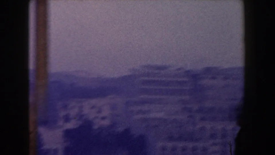 Color-shifted 8mm frame of a hazy Italian hillside town with mountains in the background.