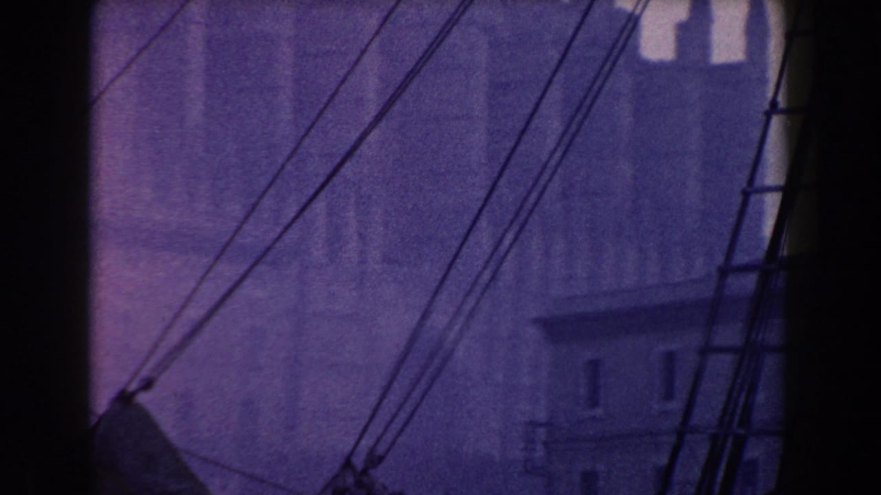1960 Italy 8mm frame with ship rigging crossing a soft view of waterfront buildings and a tall vertical structure.