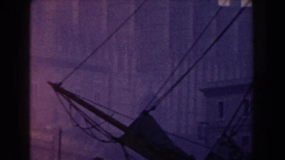 1960 Italy 8mm view of a boat boom, rigging lines, and soft-focus waterfront buildings in the background.
