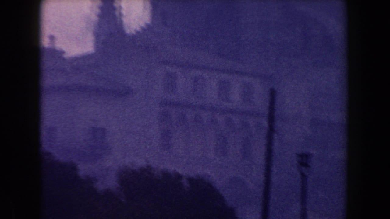 1960 Italy 8mm frame showing a misty city street, large building mass, distant spire, and soft purple color shift.