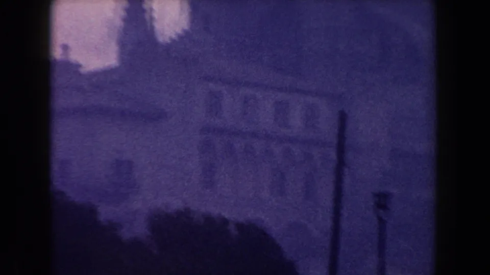 1960 Italy 8mm frame showing a misty city street, large building mass, distant spire, and soft purple color shift.