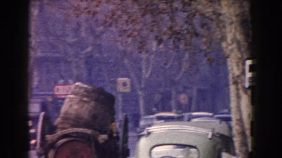 1960 Italy street view with a large barrel on a cart, rounded cars, pedestrians, trees, and visible storefront signage.