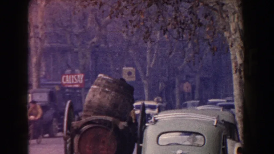 1960 Italy 8mm frame with a barrel cart in the foreground, mid-century cars, pedestrians, tree trunks, and shop signage.