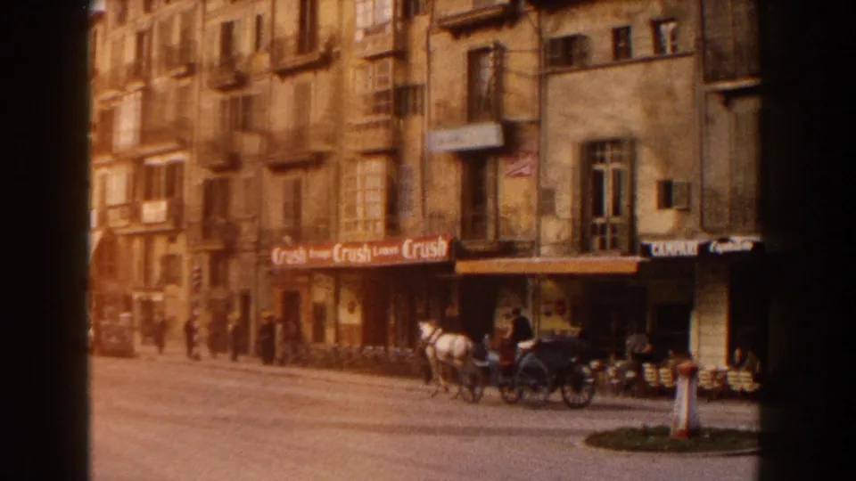 1960 Italy street scene showing weathered apartment facades, storefront signage, parked cars, pedestrians, and a horse-drawn carriage.