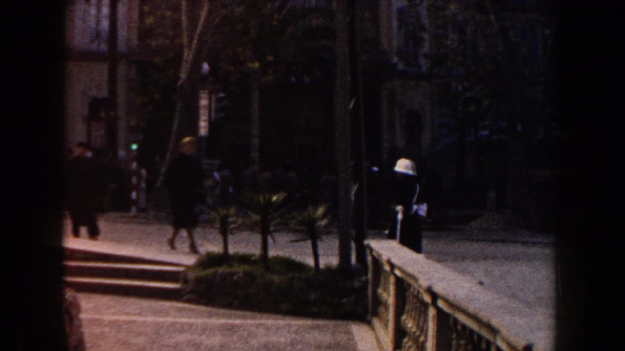 1960 Italy home-movie frame with pedestrians near a stepped walkway, railing, trees, and a shaded urban building entrance.