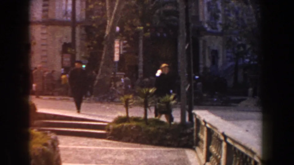 1960 Italy home-movie frame showing people walking by steps, a railing, trees, and older stone or stucco building facades.