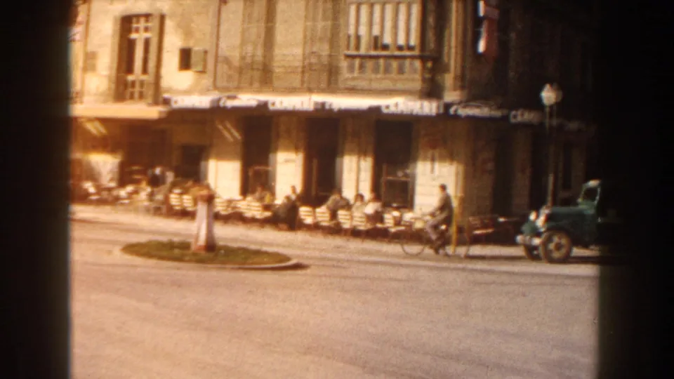 1960 Italy home-movie frame showing cafe awnings, street seating, pedestrians, bicycles, a parked vehicle, and older buildings.
