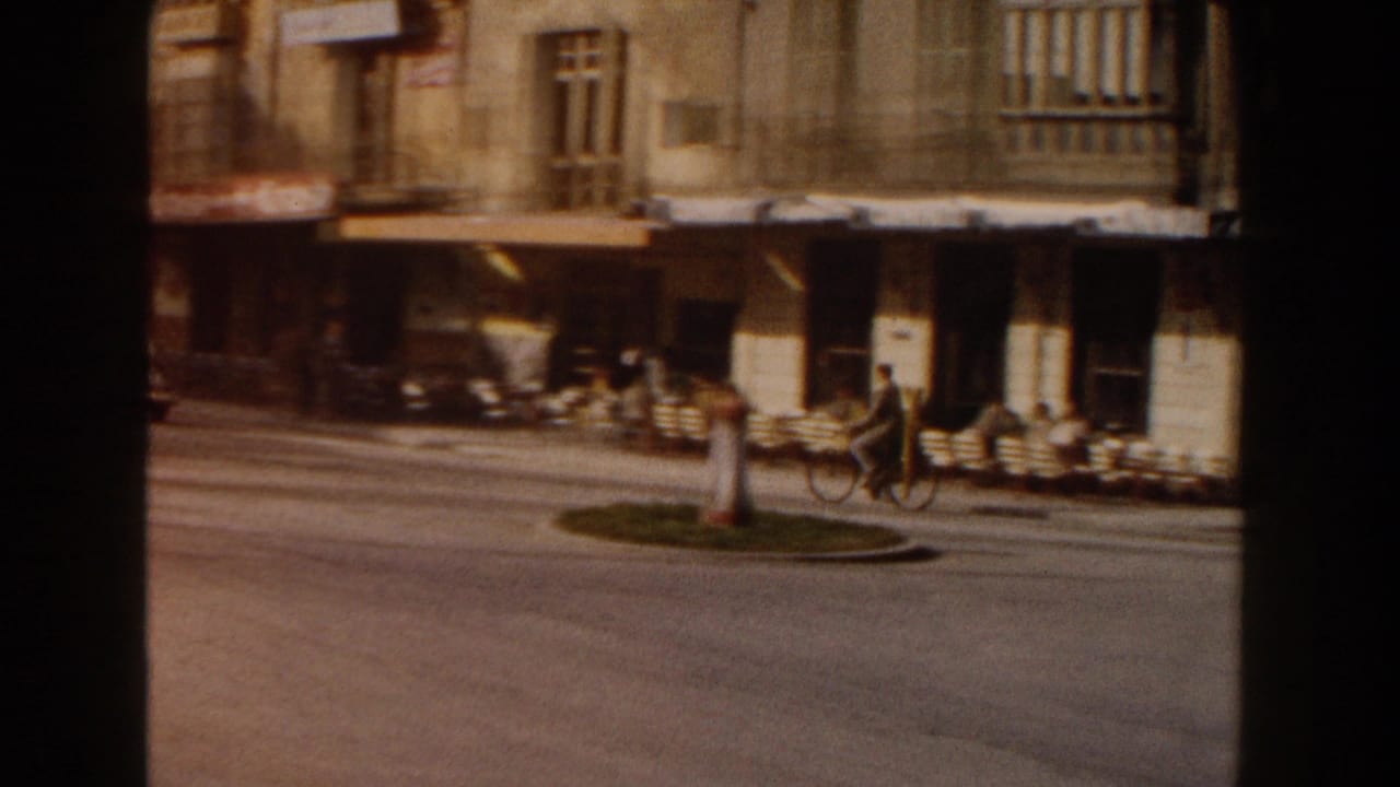 1960 Italy street view with a small roundabout, cafe tables under awnings, people by the curb, and two-wheeled traffic in soft 8mm color.