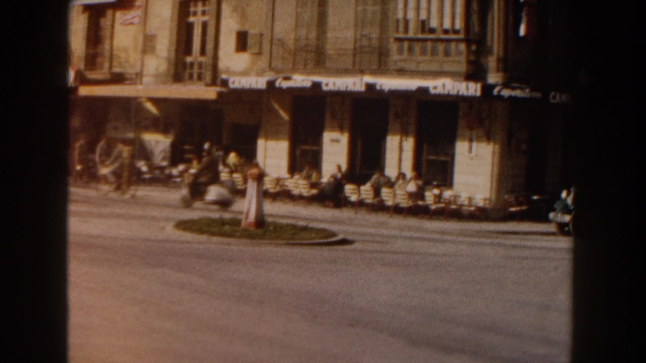1960 Italy street scene showing outdoor cafe seating, pedestrians, motorcycles or bicycles, shop awnings, and a small roundabout in the road.