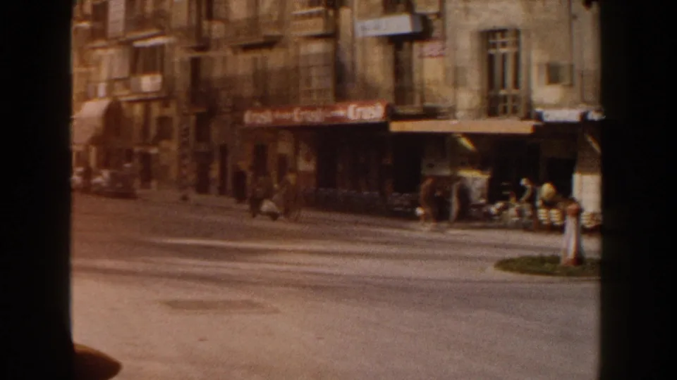 1960 Italy view across a city street toward shopfronts, pedestrians, cafe signage, a small roundabout, and passing traffic.