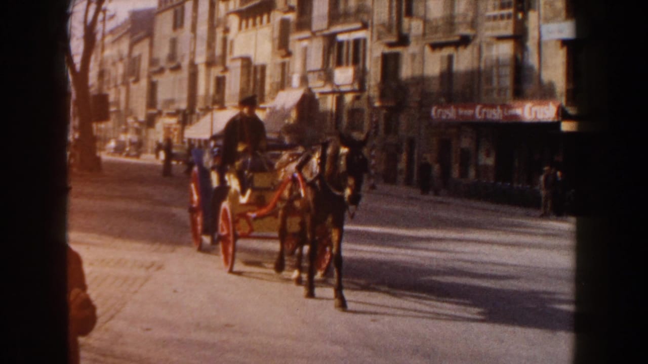 In 1960 Italy home-movie footage, a horse-drawn carriage with red wheels moves along a sunlit street past storefronts and pedestrians.