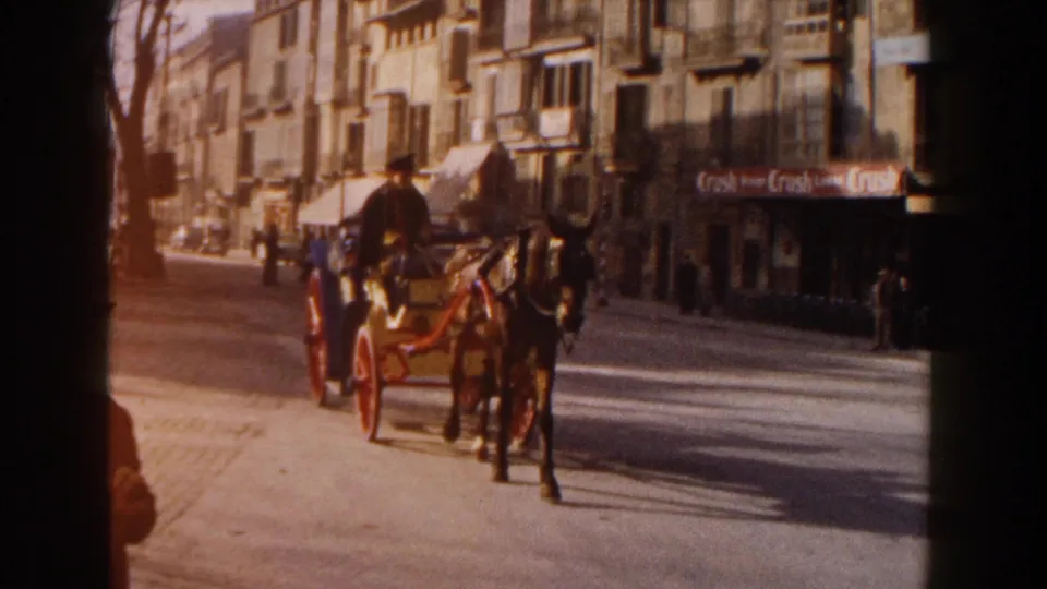 In 1960 Italy home-movie footage, a horse-drawn carriage with red wheels moves along a sunlit street past storefronts and pedestrians.