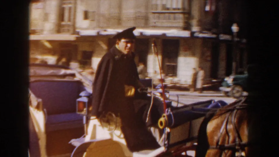 1960 Italy 8mm frame showing a caped man seated on a horse-drawn buggy beside urban buildings.