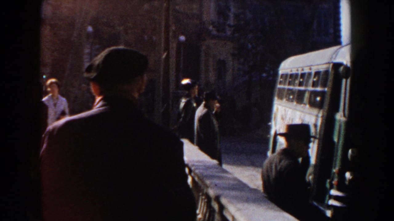 1960 home-movie frame showing pedestrians in dark clothing near a bus along a shaded Italian street or square.