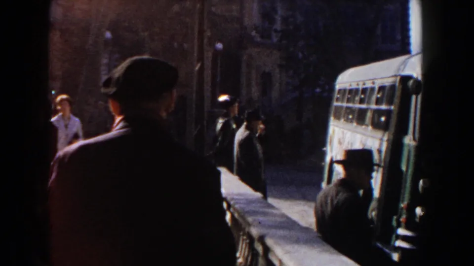 1960 home-movie frame showing pedestrians in dark clothing near a bus along a shaded Italian street or square.
