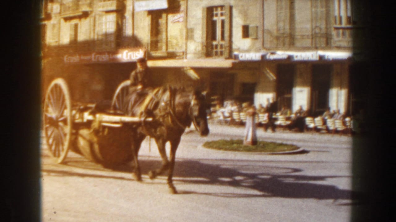 1960 Italian street scene with a horse-drawn cart crossing a sunlit square beside café awnings and outdoor tables.