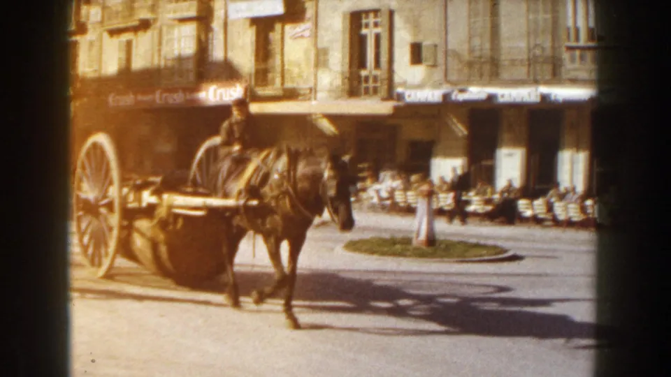 1960 Italian street scene with a horse-drawn cart crossing a sunlit square beside café awnings and outdoor tables.