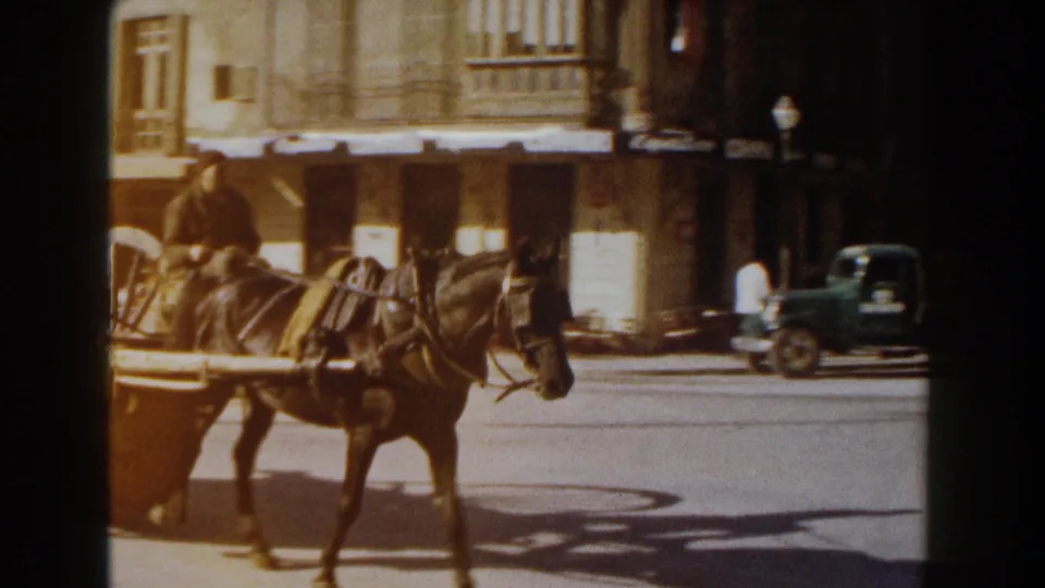 Horse-drawn cart moving through an Italian square, with café frontage, awnings, and a parked vehicle in the background.