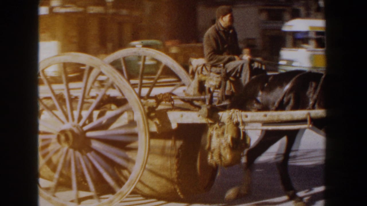 1960 Italy home-movie frame showing a horse-drawn wooden cart with a barrel and seated driver passing urban buildings in sunlight.