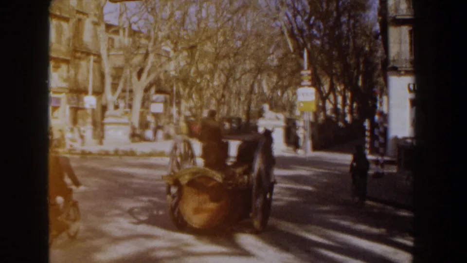 1960 Italy home-movie frame with the barrel cart moving away on a sunny street, with a cyclist, pedestrians, trees, and buildings nearby.