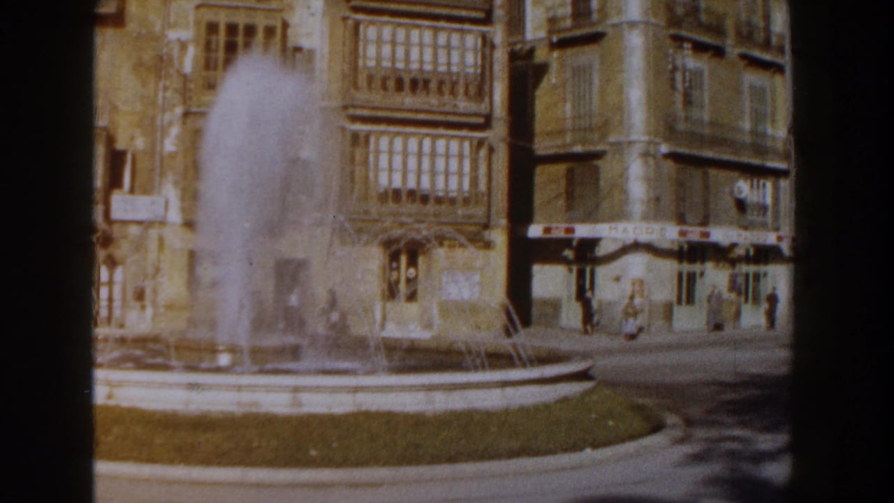 1960 Italy 8mm frame showing a plaza fountain spraying before tan masonry buildings with enclosed balconies and street-level awnings.