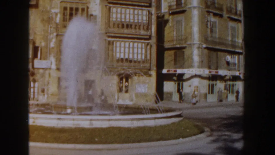 1960 Italy 8mm frame showing a plaza fountain spraying before tan masonry buildings with enclosed balconies and street-level awnings.