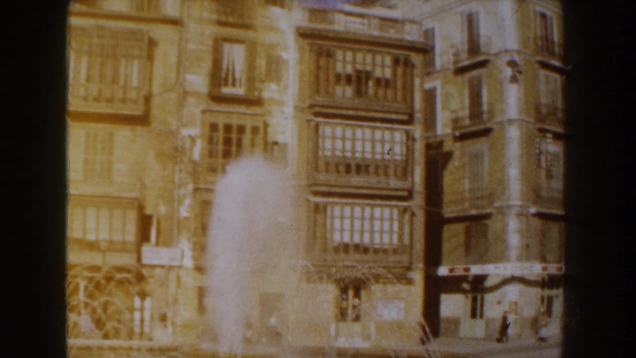 Warm 8mm view of fountain water rising in front of tan Italian building facades and enclosed balconies, with dark film borders visible.