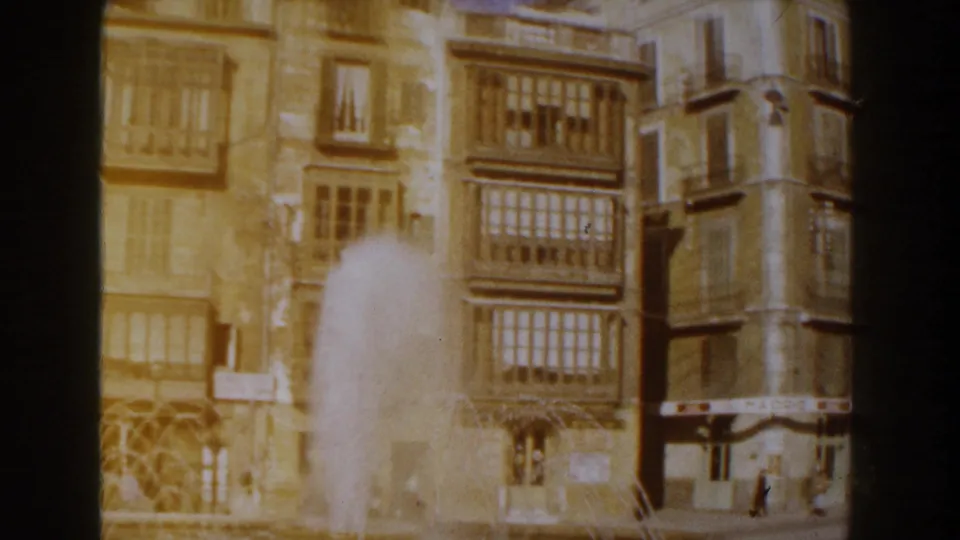 Warm 8mm view of fountain water rising in front of tan Italian building facades and enclosed balconies, with dark film borders visible.