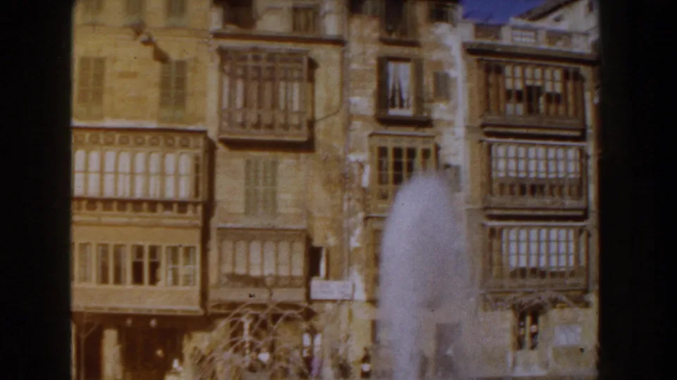 1960 Italy home-movie frame with a vertical view of tan buildings, enclosed balconies, windows, and fountain spray crossing the lower foreground.