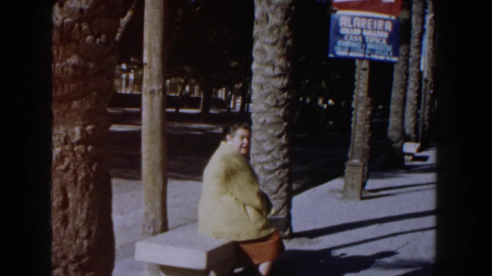 1960 Italy 8mm frame showing a woman seated near palm trees, a sidewalk, parked car edge, and a blue commercial sign in the background.