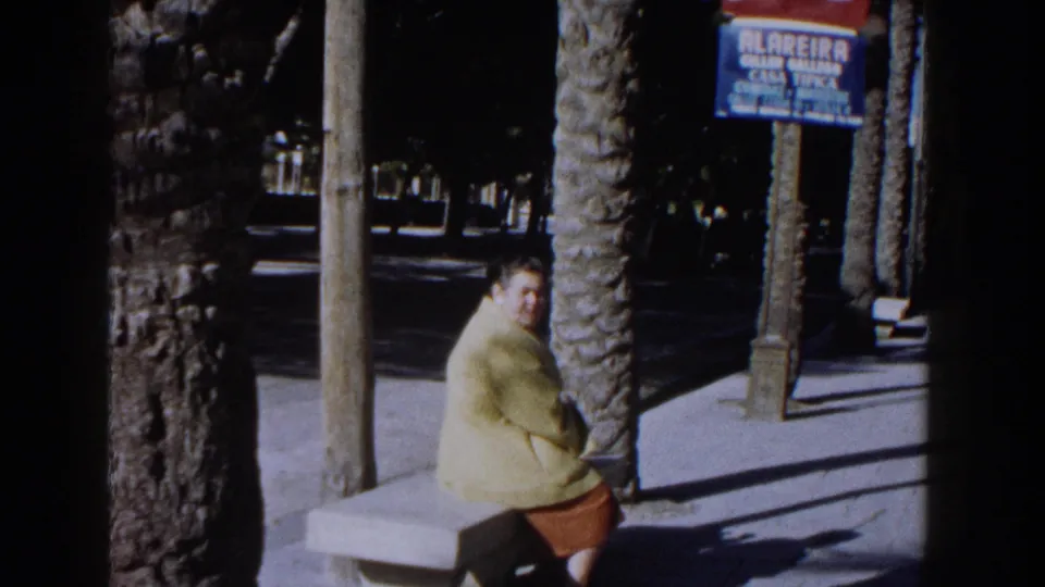 1960 Italian home-movie frame of a woman in a yellow jacket seated on a stone bench in a palm-lined square.