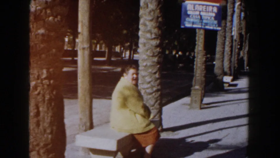 The woman smiles from a stone bench in the Italian street scene, with palm trunks, strong sun, and a blue sign visible.