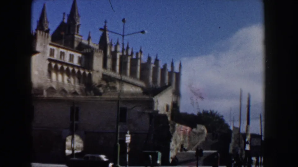 1960 Italy street view with a historic cathedral, crenellated wall, parked cars, signs, lamp posts, and small pedestrians in the distance.