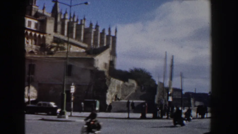 1960 Italy 8mm frame showing the cathedral above a public street with parked cars, pedestrians, lamp posts, and clouds near the horizon.