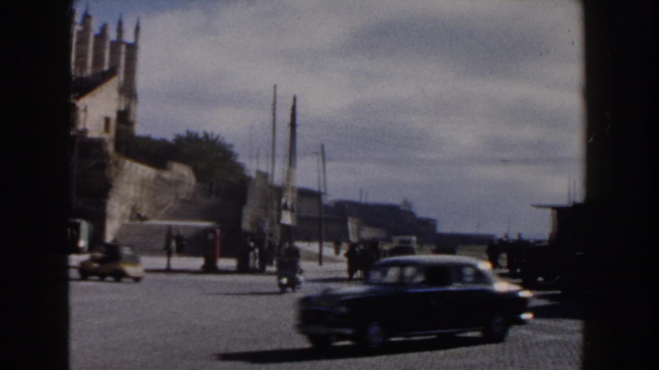 Dark car crosses a broad street near a Gothic-style building and distant pedestrians.