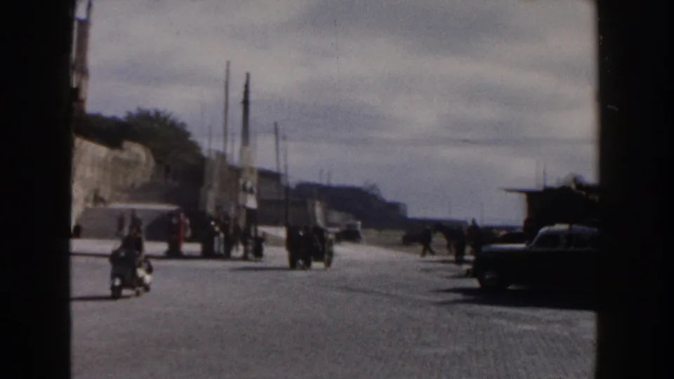 1960 Italy street scene with a motor scooter or motorcycle, pedestrians, cars, utility poles, and stone walls near historic architecture.