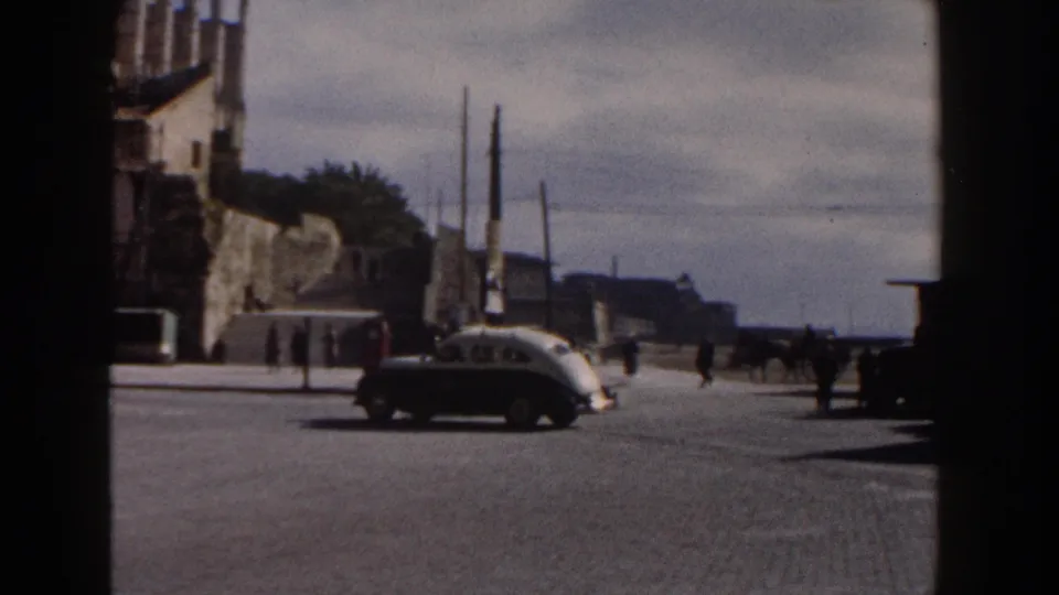 A light-colored classic car crosses a sunlit Italian street, with pedestrians, utility poles, and a Gothic-style building partly visible at left.