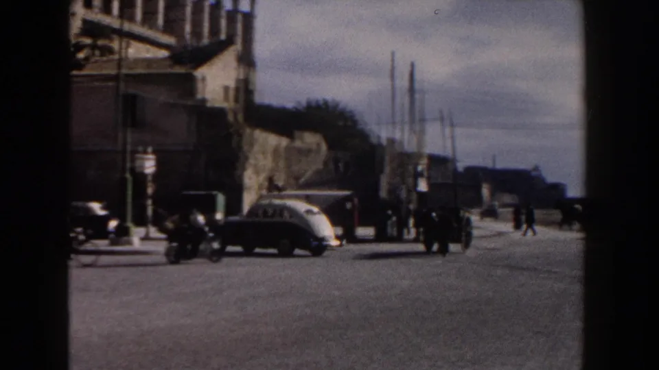 1960 Italy street view with a classic car, a motorbike, pedestrians, signs, stone architecture, and open paved roadway.