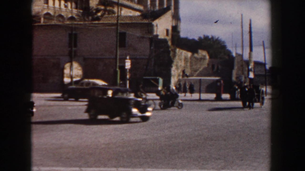 A dark classic car crosses a broad Italian street in 1960, with pedestrians, a motorbike, stone architecture, and tall poles nearby.
