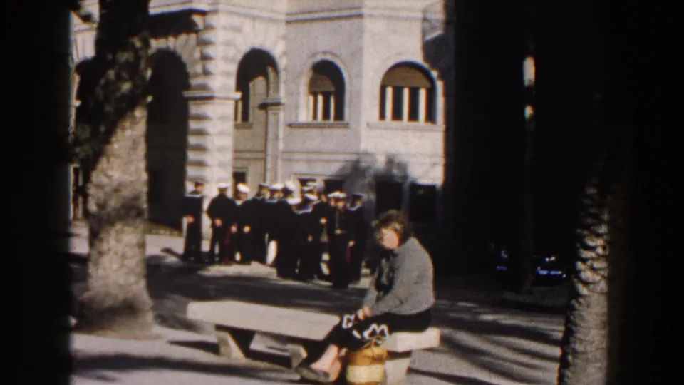 1960 Italy public street scene with a woman seated on a bench, uniformed men nearby, palm trees, steps, and arched stone architecture.