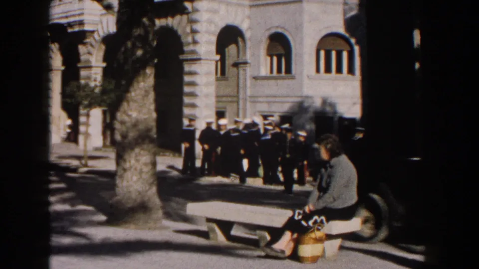 A 1960 Italian street frame shows a woman on a bench, uniformed men gathered nearby, palm trees, a dark car, and arched stone windows.