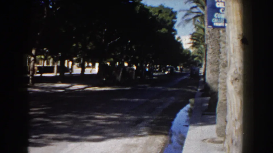 1960 Italy 8mm frame with a tree-lined street, bright curb, distant vehicles, a blue sign, and a small cyclist or figure in the shade.