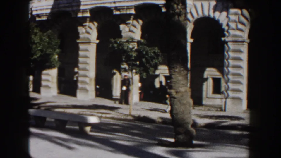1960 Italy 8mm frame showing a stone facade with arched colonnades, a small tree, bench, and pedestrians passing in shade.
