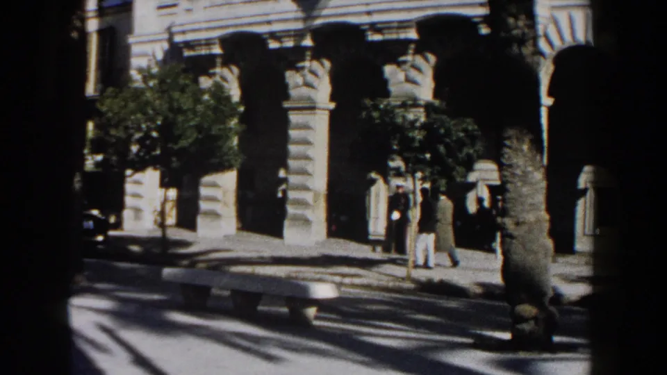 1960 Italy frame with people walking near arched stone colonnades, a palm trunk, bench, and strong street shadows.