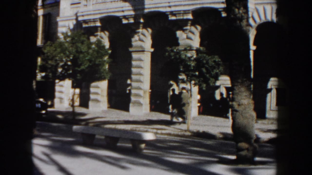 1960 Italy street view with pedestrians near an arched beige stone facade, trees, a bench, and strong pavement shadows.