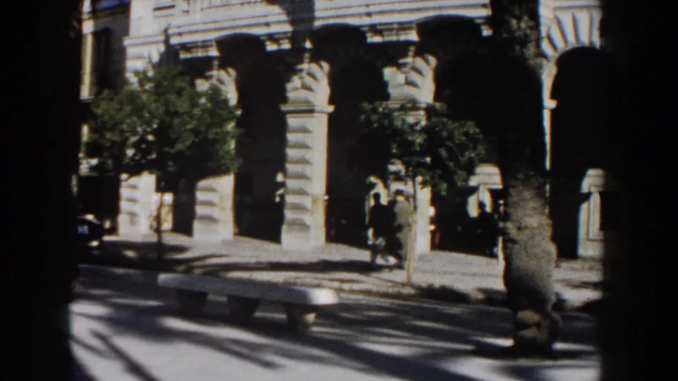1960 Italy street view with pedestrians near an arched beige stone facade, trees, a bench, and strong pavement shadows.