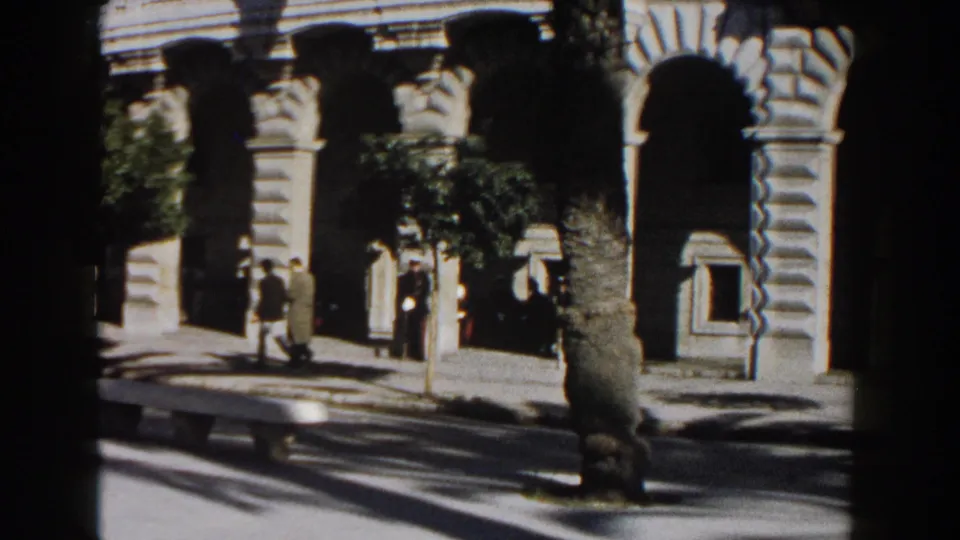 Across-street 1960 Italy view of pedestrians beside a beige arched building, with trees and high-contrast sunlight.
