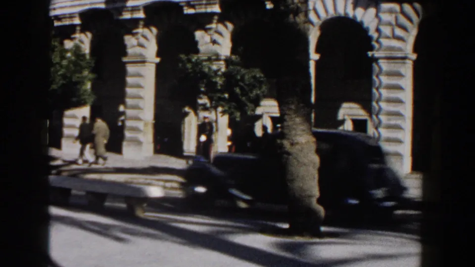 A dark car moves across the foreground of a 1960 Italy street scene with pedestrians and an ornate stone building behind it.